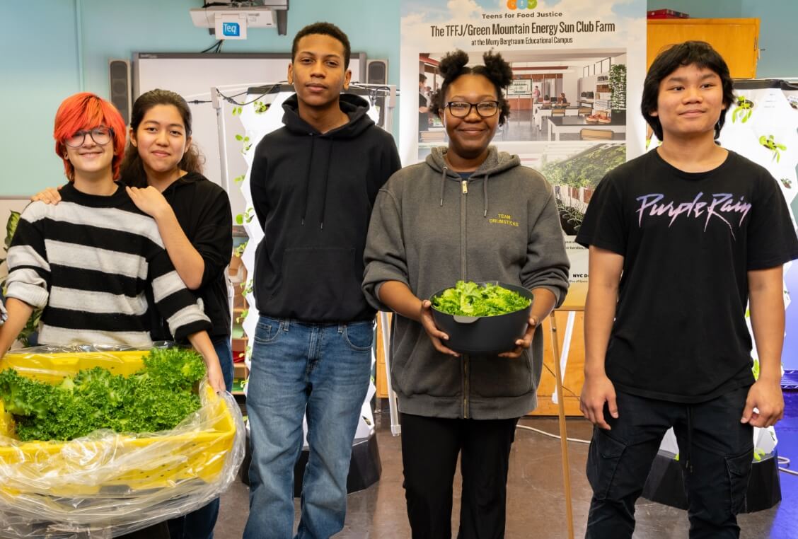 Five young people stand indoors holding harvested lettuce, with a poster about a school farm project displayed in the background.