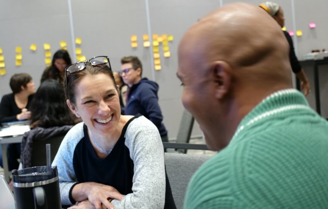 Two Springpoint Fellows sit across from each other at a table, smiling and talking in a room with other people and sticky notes on the wall in the background.